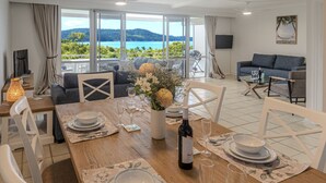 A dining area with a table set for four, a bottle of wine, and a view of the sea through large windows.