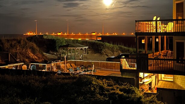 A beachfront property with a wooden deck, a covered seating area, and a view of the ocean and distant city lights. - Spacious ocean front house with spectacular views in Nags Head with Elevator (Nags Head)