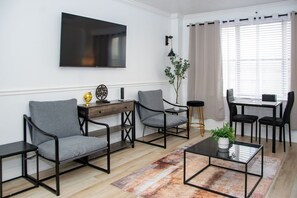 Ein modernes Wohnzimmer mit Flachbildfernseher, zwei grauen Sesseln, einem Couchtisch, einem Schreibtisch und einem Essbereich mit Stühlen.