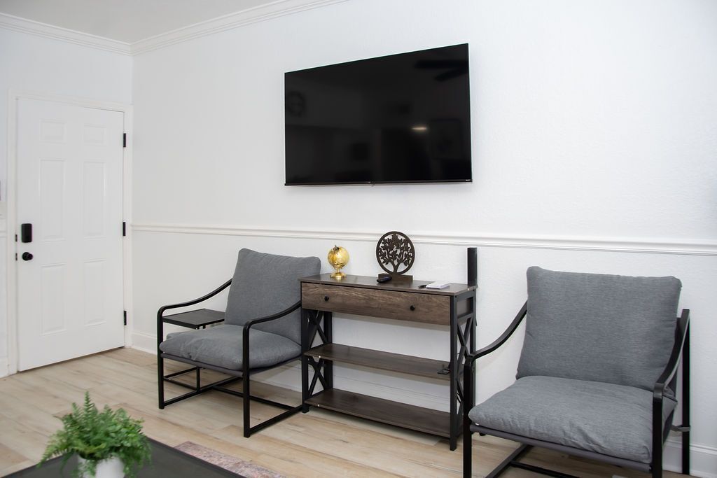 Une pièce avec une porte blanche, un téléviseur noir fixé au mur et deux fauteuils gris, séparés par un bureau en bois.