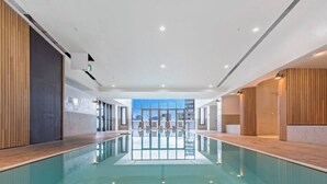 A modern indoor swimming pool with clear glass walls, a central lane, and seating areas. - 3 Bedroom CBD Apartment with Pool, Gym & Spa with Gorgeous City Views (Melbourne)