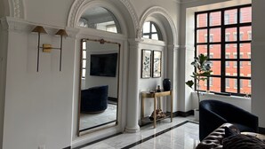 A spacious room with a large mirror, a blue chair, a small table, and a view of a red building through a window. - Luxury Downtown Indy • Walk to NCAA Final Four • On American Legion Mall (Indianapolis)