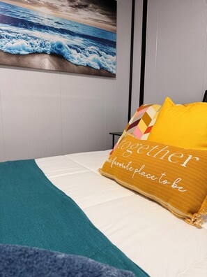 A bed with a teal throw blanket, a yellow pillow with embroidered text, and a wall-mounted picture of ocean waves. - Beautiful 2-Bedroom modular home in the tranquil country side of Jobos Isabela. (Isabela)