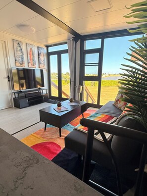 A modern living room with a television, a coffee table, and a sofa. There are large windows offering a view of a vineyard. - Beautiful 2-Bedroom modular home in the tranquil country side of Jobos Isabela. (Isabela)
