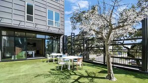 Ein modernes Gebäude mit großen Fenstern, einer Terrasse mit Tischen und Stühlen und einem Baum im Vordergrund.