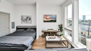 Ein modernes Schlafzimmer mit einem Bett, einem Nachttisch mit Pflanze, einem Fenster mit Stadtblick und einem Bild an der Wand.