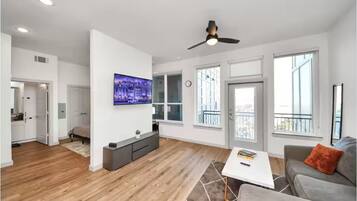A spacious living area with a ceiling fan, a flat-screen TV, and a large window.