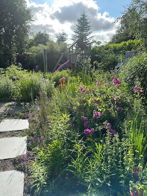 A garden with a wooden structure, a paved path, and various plants. - Vacation home for 8 people with natural garden, fireplace & lots of space, near the North Sea (Wesselburen)