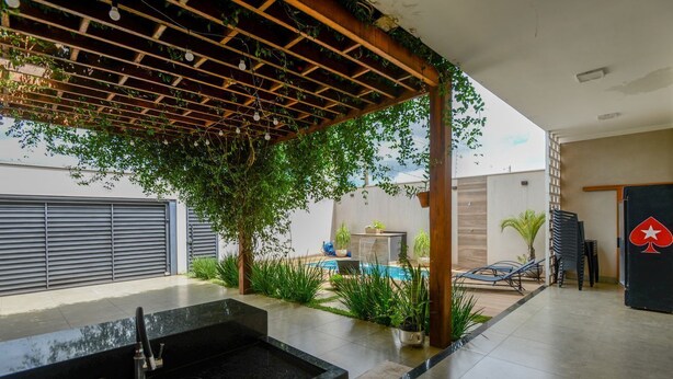 Een moderne buitenkeuken met een inbouwspoelbak, een pergola met hangende lampen en een zwembad op de achtergrond.