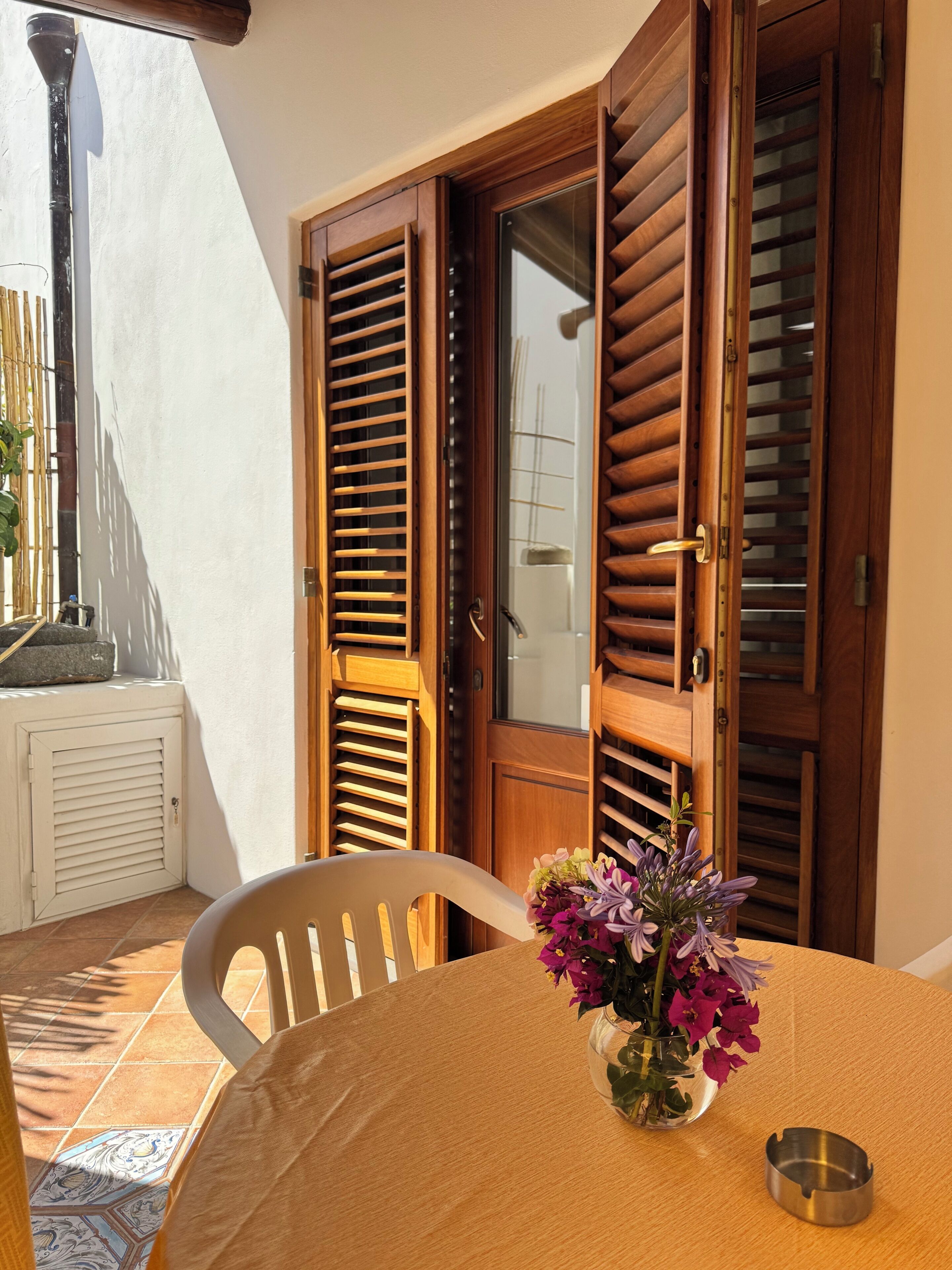 Une pièce avec une porte en bois, une chaise blanche, une table ornée d’un vase de fleurs et un sol carrelé.