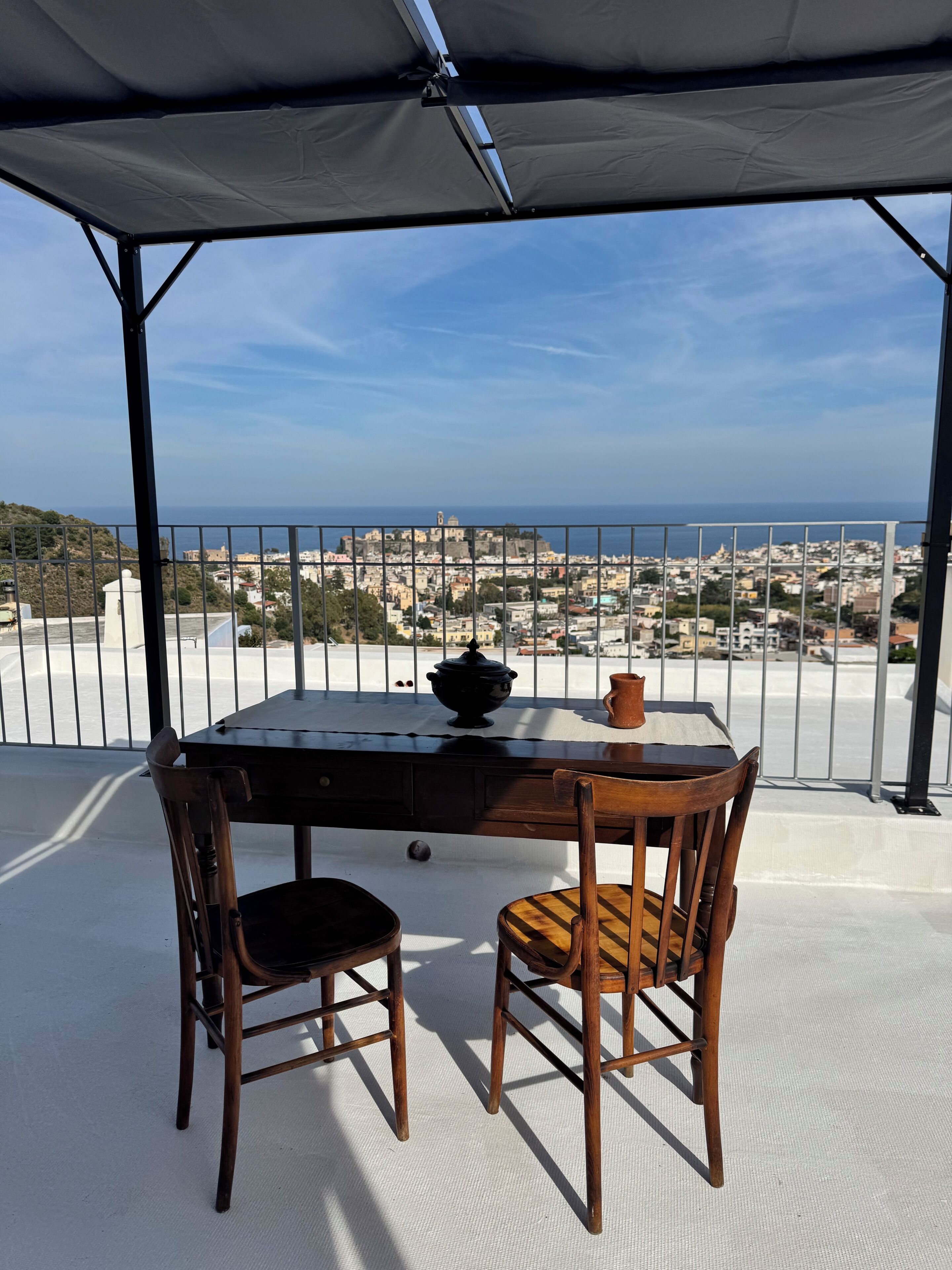 A covered outdoor seating area with a table and chairs, overlooking a coastal cityscape.