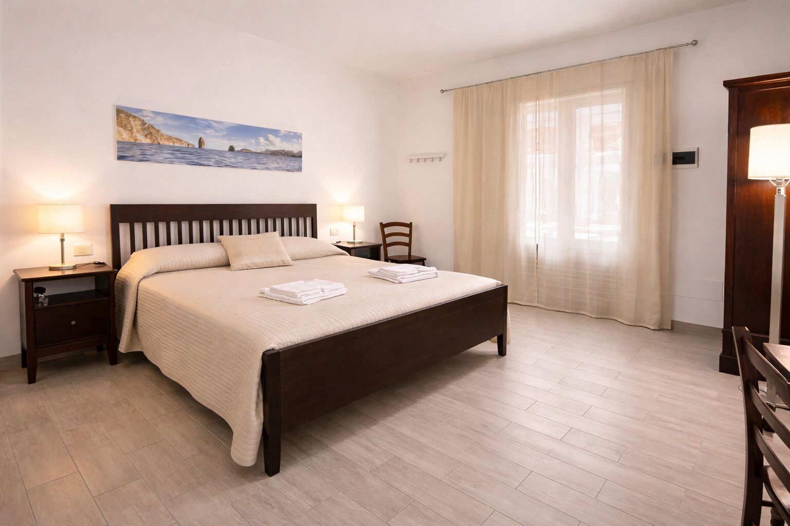 Un lit bien fait, avec une tête de lit, deux tables de chevet avec des lampes, une chaise et une grande fenêtre avec des rideaux.