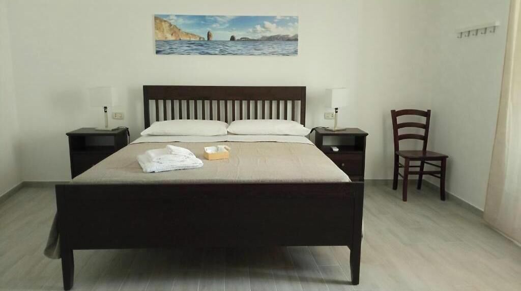 A bed with a wooden headboard, two bedside tables, a chair, and a painting of a coastal scene.