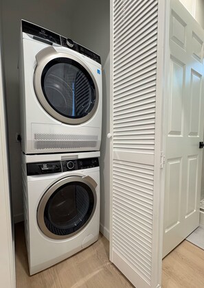 A stacked washer and dryer unit in a room with a wooden floor and a white door. - Sunny Cypress Retreat - 1BR + Bonus Bed - New ADU (Cypress)