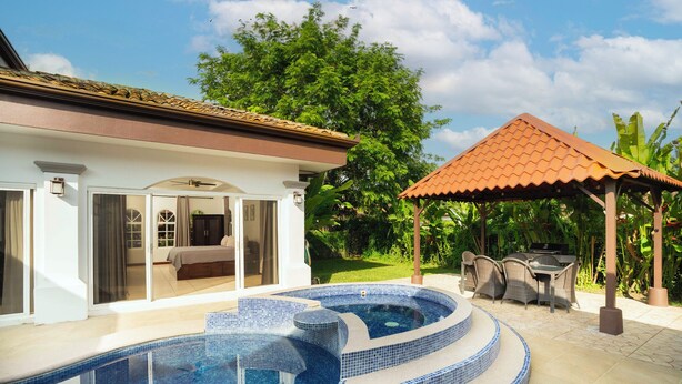 Une piscine avec un bord carrelé, un patio couvert et une maison avec un toit en tuiles.