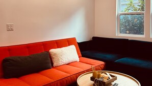 A modern living room with a red sofa, a dark blue sofa, a round wooden coffee table, and a large window with a view of greenery. - Boutique 1BR Private Unit with AC, Silicon Valley, 5 min Santana Row, fast WiFi (San Jose)