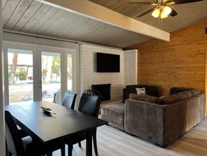 A modern living room with a wooden ceiling, a flat-screen TV mounted on a brick wall, a dark wooden table, and a gray sofa. - 2-bedroom house in charming Rancho Mirage with AC (Rancho Mirage)