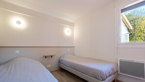 A small, well-lit room with two beds, a window with a view of trees, and a built-in heater. - Gite 6/8 pers. PREMIUM - Village des Bruguières (LA CANOURGUE)