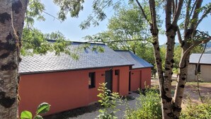 A small building with a red exterior and a grey roof, surrounded by trees. - Gite 6/8 pers. PREMIUM - Village des Bruguières (LA CANOURGUE)