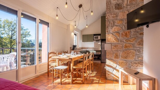 A modern kitchen with a stone wall, a dining area with wooden furniture, and a balcony with chairs. - Gite 6/8 pers. PREMIUM - Village des Bruguières (LA CANOURGUE)