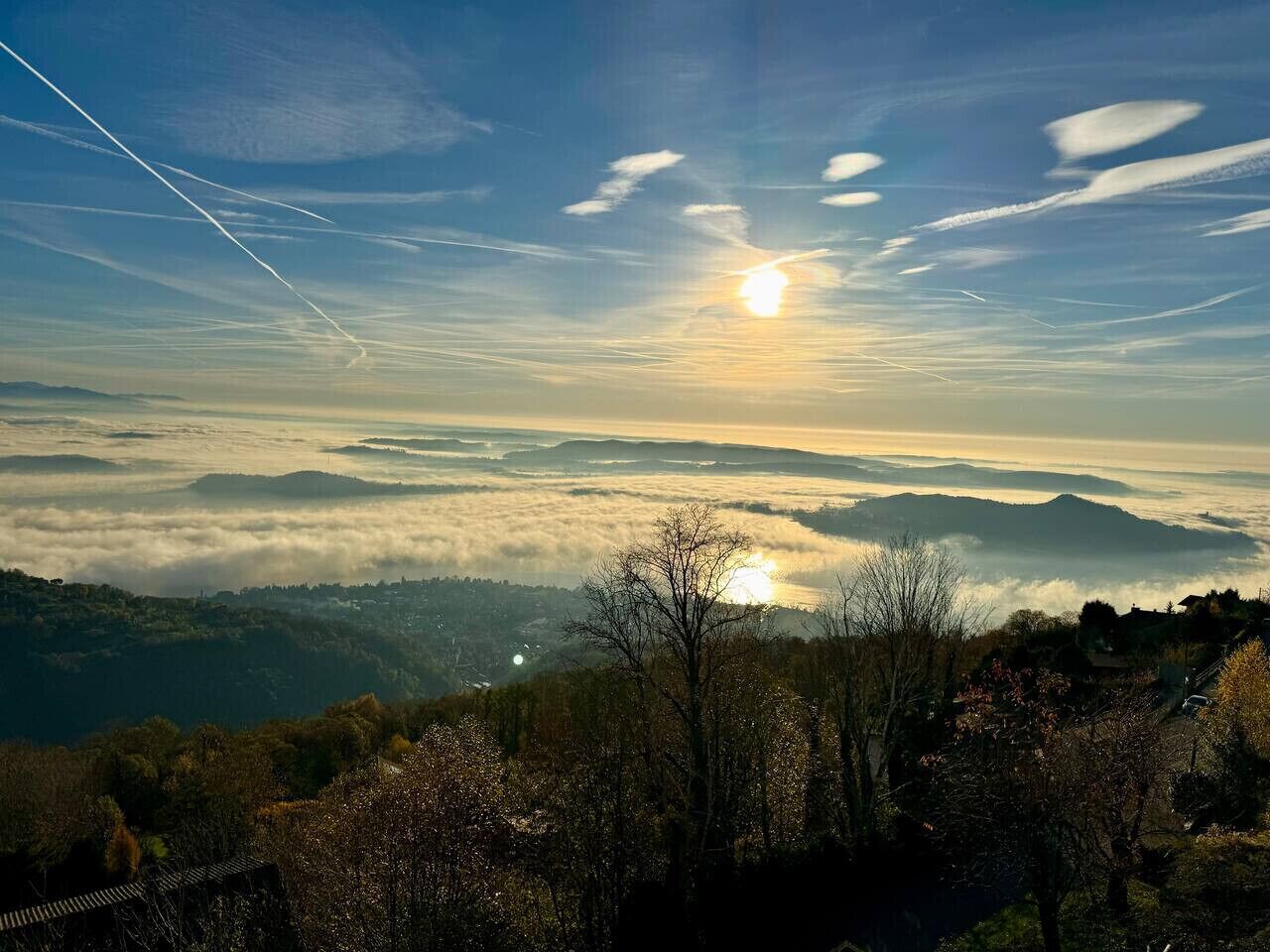 A panoramic view of a mountainous landscape with a sunrise or sunset, featuring a clear sky, distant mountains, and a foreground with trees and a structure.