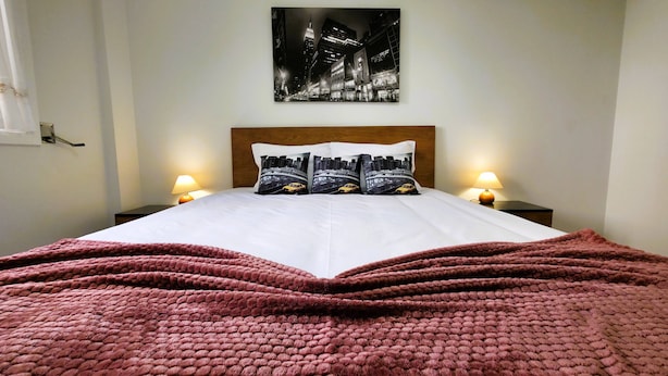 Ein ordentlich bezogenes Bett mit roter Decke, zwei Nachttischlampen und einem Stadtbild-Wandbild.