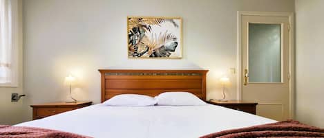 Ein Hotelzimmer mit einem großen Bett, zwei Nachttischlampen, einem hölzernen Kopfteil, einem gerahmten Kunstwerk an der Wand und einer Tür mit satiniertem Glas.