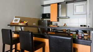 Uma cozinha compacta com micro-ondas, fogão e armários laranja. Há cadeiras pretas e uma pequena mesa com uma cesta de frutas.