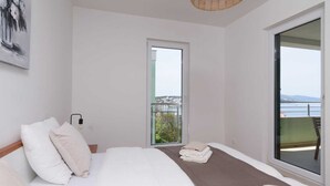 Quarto com cama, almofadas, mesa de cabeceira, luminária, janela e vista para o mar.