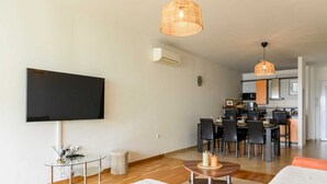 Uma sala de estar moderna com uma TV de tela plana, uma área de jantar com cadeiras, uma cozinha ao fundo e uma janela grande.