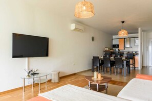 Ein modernes Wohnzimmer mit Flachbildfernseher, einem Essbereich mit Stühlen, einer Küche im Hintergrund und einem großen Fenster.