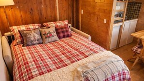 Un lit avec une couvre-lit à carreaux et des oreillers, un mur en bois, une petite table avec une chaise, et un coin cuisine avec des placards et un dosseret carrelé.