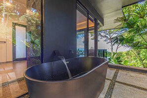 Un baño moderno con una bañera grande, una puerta de cristal que da a un balcón con vistas a zonas verdes.