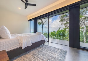 A modern bedroom with a bed, a ceiling fan, and a large window offering a view of greenery. - Brand-New Ocean View Home at a Special Rate (Piedra Amarilla)