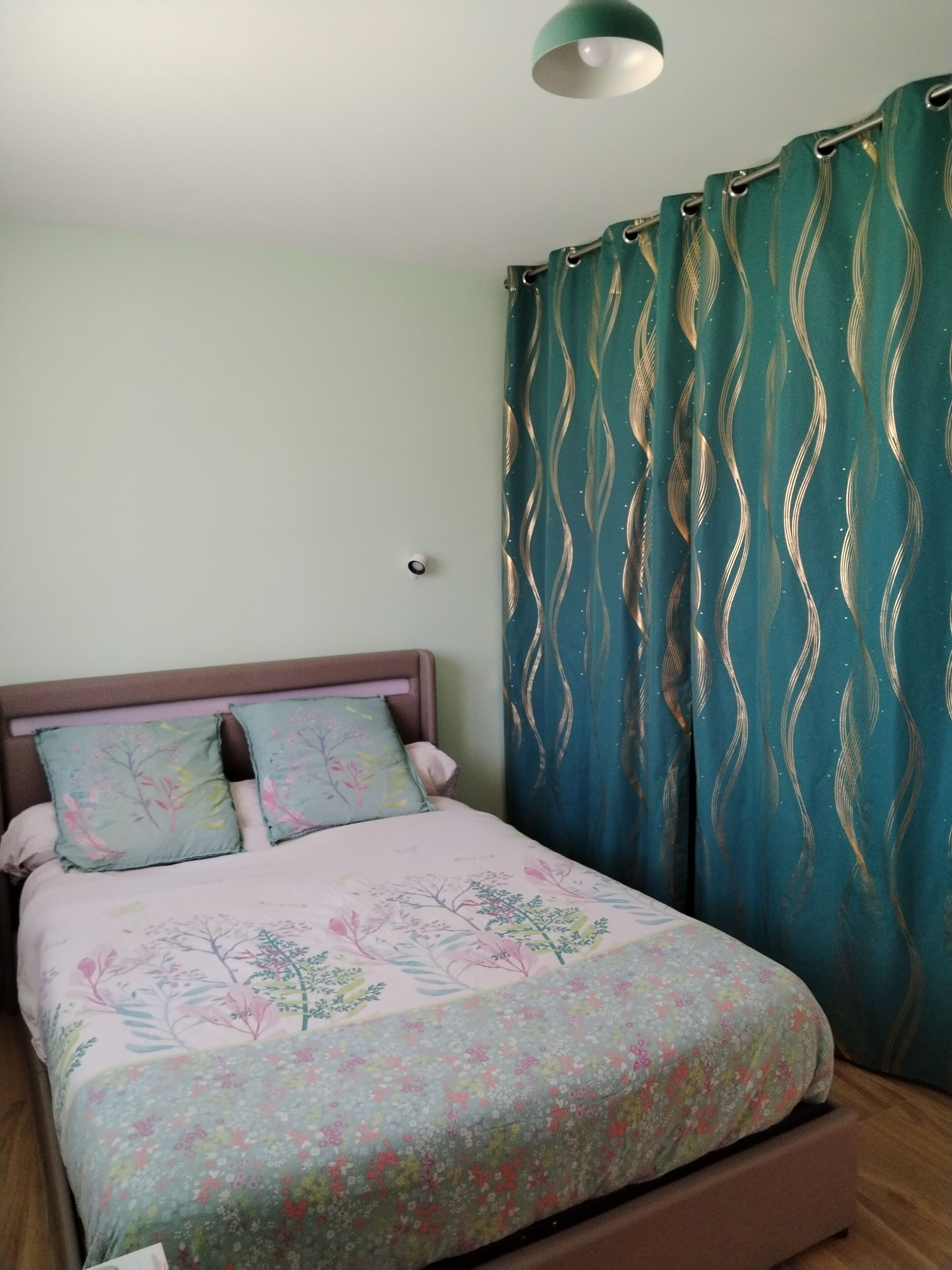 A bedroom with a bed, two pillows, a headboard, a nightstand, a wall-mounted light, and a curtain.