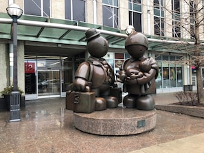 Une sculpture représentant deux personnages, l’un tenant une valise et l’autre un bébé, devant un bâtiment doté d’une entrée en verre et d’une enseigne indiquant « 18 Yonge ».