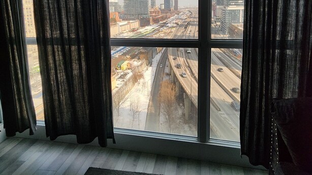 A room with a large window, a black ottoman, and a view of a cityscape with buildings and a highway. - 2-bedroom apartment in amazing Toronto with cool AC breeze (Toronto)