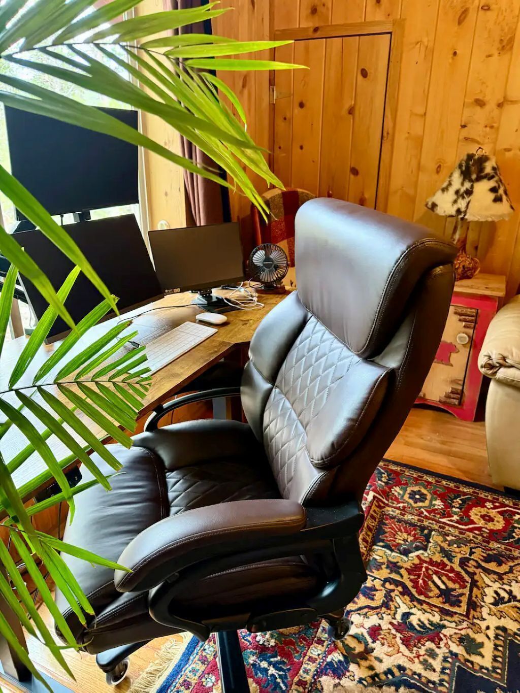 Uma cadeira de escritório de couro, uma mesa com um computador, um ventilador e uma planta.