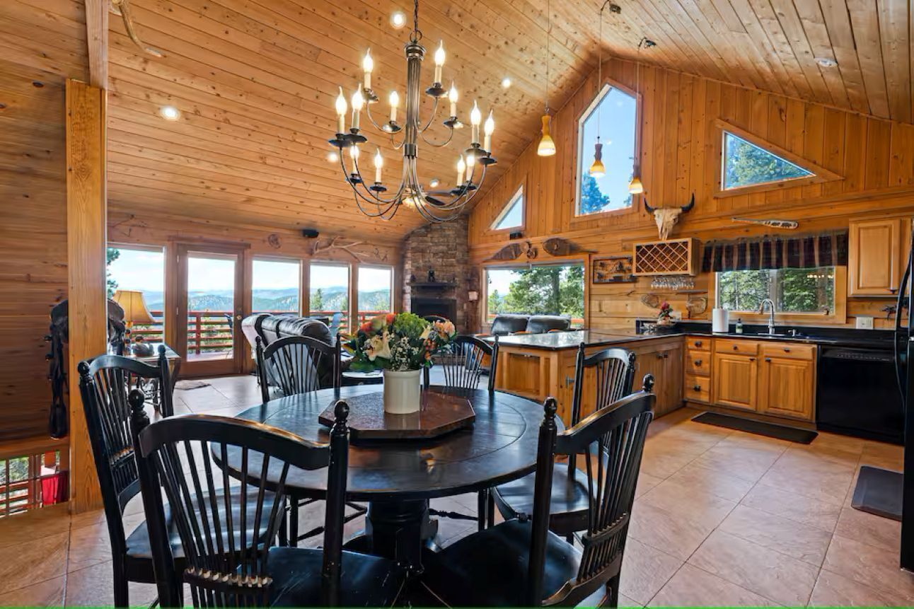 Interno di una baita in legno con tavolo da pranzo, sedie, un lampadario, angolo cottura e ampie finestre.