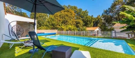 A backyard with a swimming pool, a lounge chair, a sun umbrella, and a small outdoor seating area.