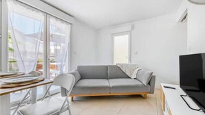 Een moderne woonkamer met een grijze bank, een eettafel met stoelen, een televisie en een groot raam met gordijnen.
