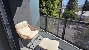 A balcony with a wicker chair and a small ottoman, a metal railing, and a view of trees and buildings.