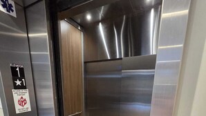 An elevator interior with a metallic finish, a door with a glass panel, and a floor with a herringbone pattern.