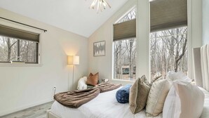 Un dormitorio con una cama grande, una lámpara de noche, una ventana con persianas y vistas a árboles fuera.