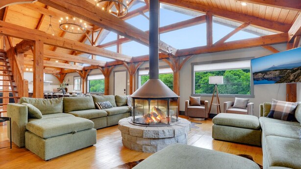 A spacious living room with a stone fireplace, wooden beams, and a large window overlooking greenery. - Pickleball, Sauna, Hot Tub, Cold Plunge, Game Room, Pond, Mini Golf (Broad Top City)