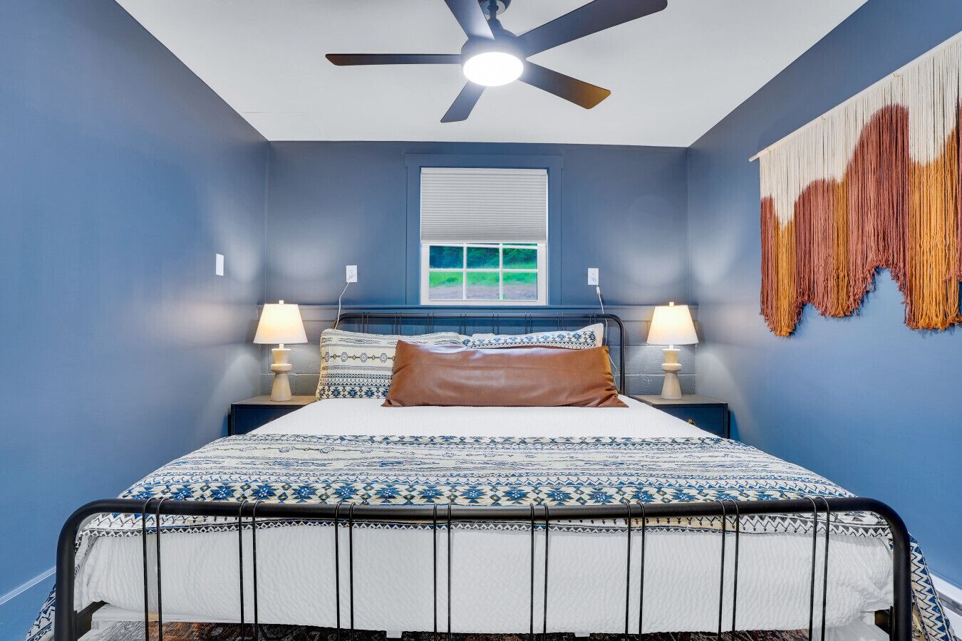 A bedroom with a bed, a ceiling fan, two bedside lamps, a window, and a wall art piece.