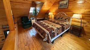 Un dormitorio acogedor en el ático con techo inclinado de madera, una cama con ropa de cama estampada, una silla verde y una mesita con lámpara.