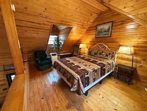 Een knus slaapkamer op zolder met een houten schuine dak, een bed met beddengoed met een patroon, een groene stoel en een kleine tafel met een lamp.