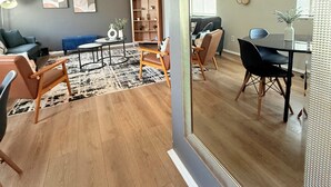A modern living room with a large mirror, wooden flooring, a blue sofa, a coffee table, and a dining area with a glass table and chairs. - Hot Tub Oasis Close to Airport 2Bd (Charlotte)