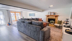 Uma sala de estar moderna com um sofá cinza, lareira e um espaço de jantar com mesa e cadeiras brancas.